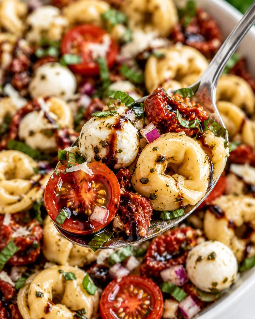 Close-up of Greek chicken orzo lifted on a stainless-steel spoon from a cream enameled Dutch oven on a bright white marble surface, showing tender seared chicken, creamy orzo, tomatoes, olives, chickpeas, spinach, and crumbled feta.
