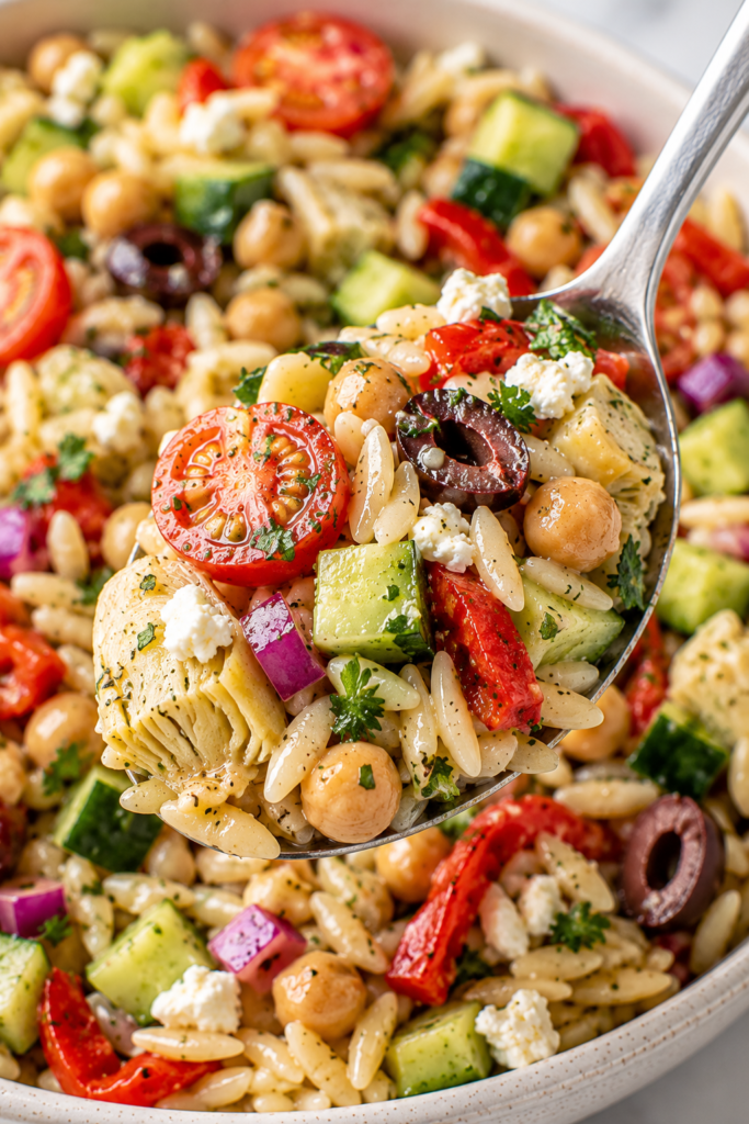 Vertical close-up of Mediterranean orzo salad lifted on a stainless steel spoon from a modern speckled ceramic bowl on a bright white marble surface, with chickpeas, cherry tomatoes, cucumber, roasted red peppers, artichoke hearts, olives, feta, parsley, and glossy dressing.
