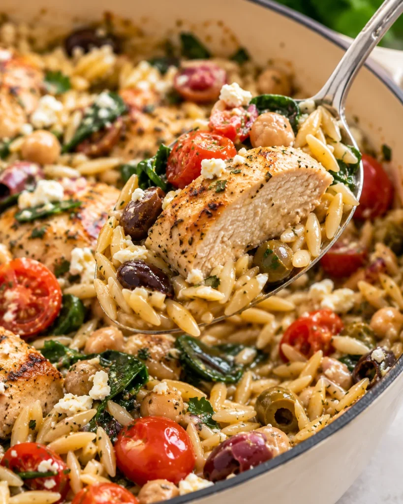 Close-up of Greek chicken orzo lifted on a stainless-steel spoon from a cream enameled Dutch oven on a bright white marble surface, showing tender seared chicken, creamy orzo, tomatoes, olives, chickpeas, spinach, and crumbled feta.