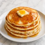 A stack of golden homemade pancakes on a white plate is topped with melting butter and glossy amber syrup on a bright white marble surface.