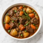 White ceramic bowl of slow cooker beef stew with tender beef, carrots, potatoes, peas, onions, rosemary, and bay leaves in glossy brown gravy on a white marble background.