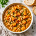 White ceramic bowl of hearty butternut squash and white bean stew with parsley, black pepper, and red pepper flakes on a white marble surface, with bread slices nearby.