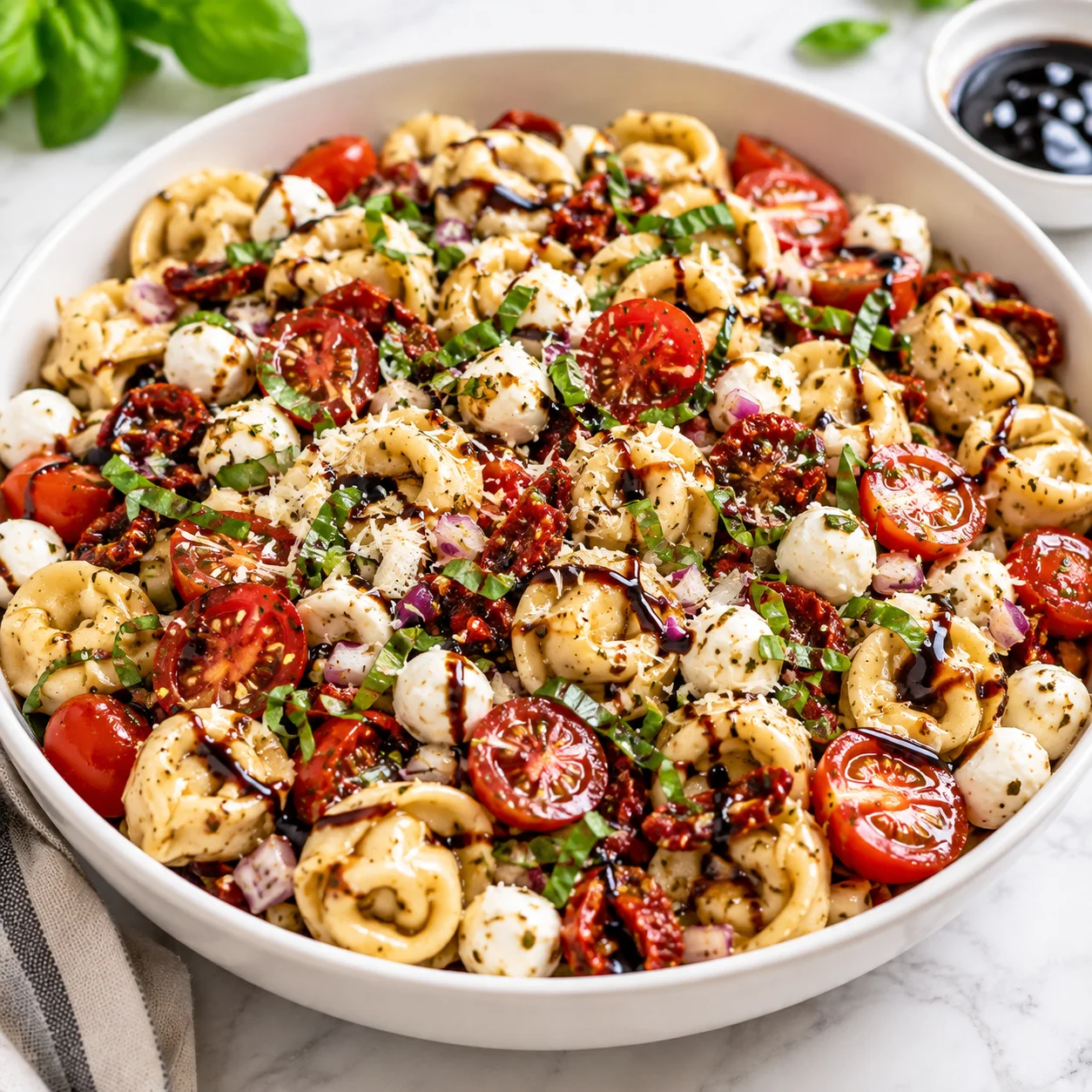 Caprese tortellini pasta salad served in a modern white ceramic bowl on a bright white marble surface, with mozzarella pearls, cherry tomatoes, sun-dried tomatoes, basil, Parmesan, red onion, and balsamic glaze.