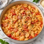 Square main recipe photograph of creamy cherry tomato shell pasta in a modern white enameled pot on a bright white marble surface, with halved cherry tomatoes, Parmesan, herbs, cracked pepper, and red pepper flakes.