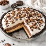 No-bake peanut butter pie in a clear glass pie dish on a bright white marble surface, with an Oreo cookie crust, peanut butter cream, chocolate pudding layer, whipped topping, and cookie crumbs showing creamy layered texture.