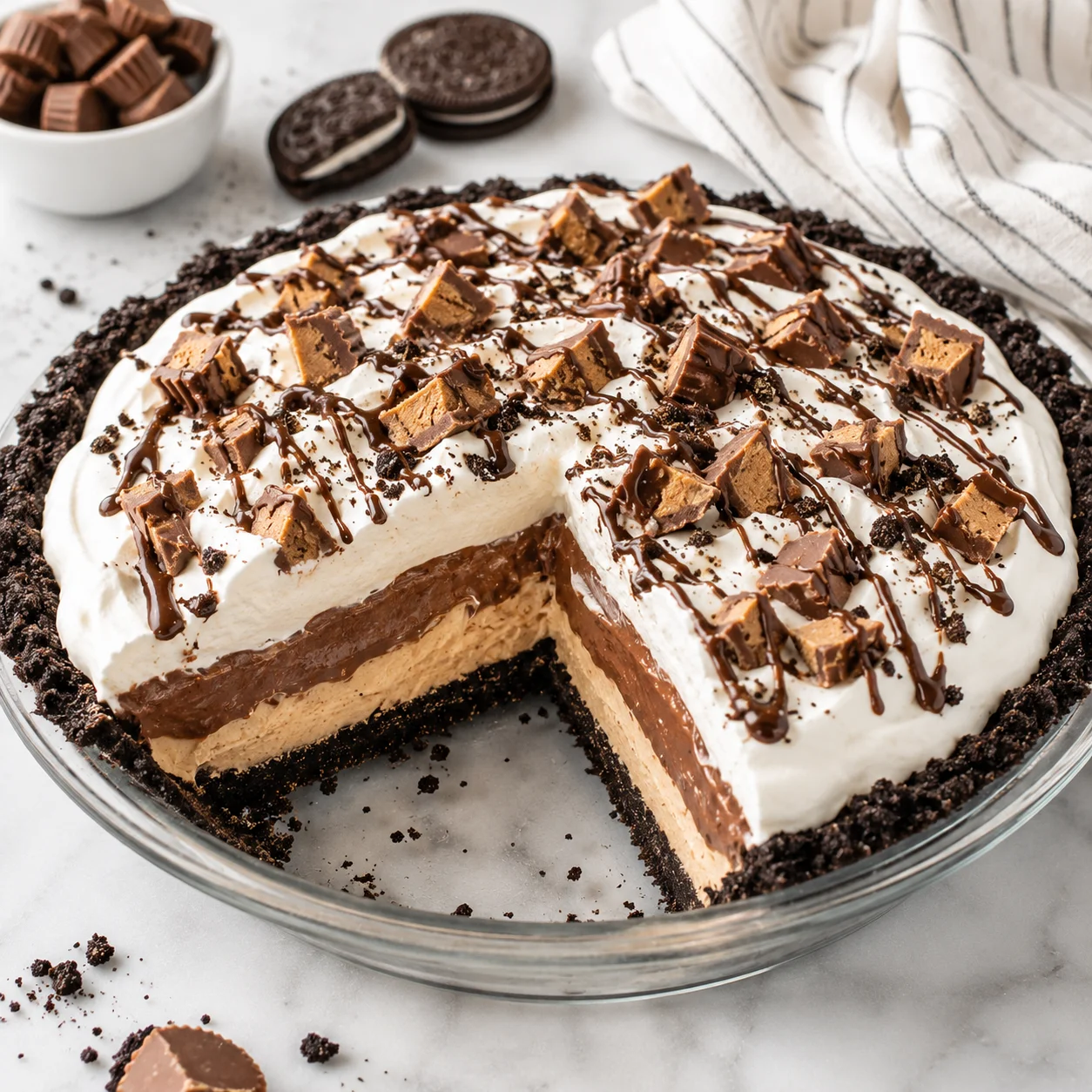 No-bake peanut butter pie in a clear glass pie dish on a bright white marble surface, with an Oreo cookie crust, peanut butter cream, chocolate pudding layer, whipped topping, and cookie crumbs showing creamy layered texture.