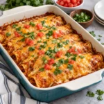 Cheesy baked chicken enchiladas in a modern ceramic baking dish on a white marble setting, topped with melted cheese, red enchilada sauce, diced tomatoes, and cilantro.