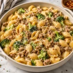 Square main recipe photograph of creamy sausage and broccoli shell pasta in a modern light ceramic pan on a bright white marble surface, topped with Parmesan, red pepper flakes, and cracked black pepper.