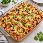 Easy Chicken Burrito Casserole baked in a clear glass dish on a bright white marble surface, with melted cheese, shredded chicken, rice, black beans, corn, red peppers, and fresh green garnish visible across the top.