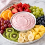 A strawberry dipped into creamy pink fruit dip being lifted from a white ceramic bowl, surrounded by fresh fruit on a bright white marble surface, showing the thick whipped texture of the dip.