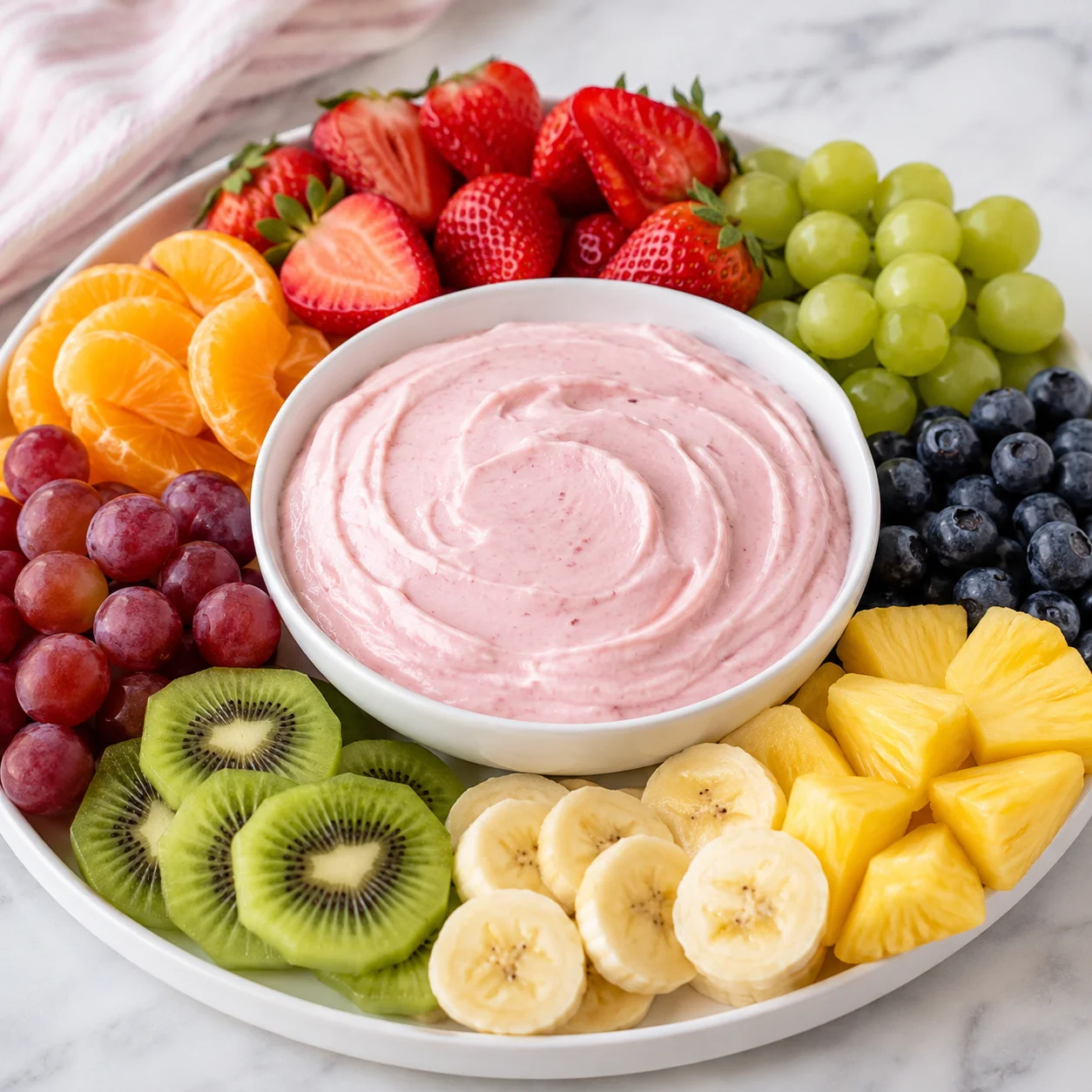 A strawberry dipped into creamy pink fruit dip being lifted from a white ceramic bowl, surrounded by fresh fruit on a bright white marble surface, showing the thick whipped texture of the dip.
