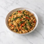 Spicy Thai basil chicken fried rice with golden chicken pieces, red chilies, and wilted basil in a speckled ceramic bowl on a clean white marble surface.